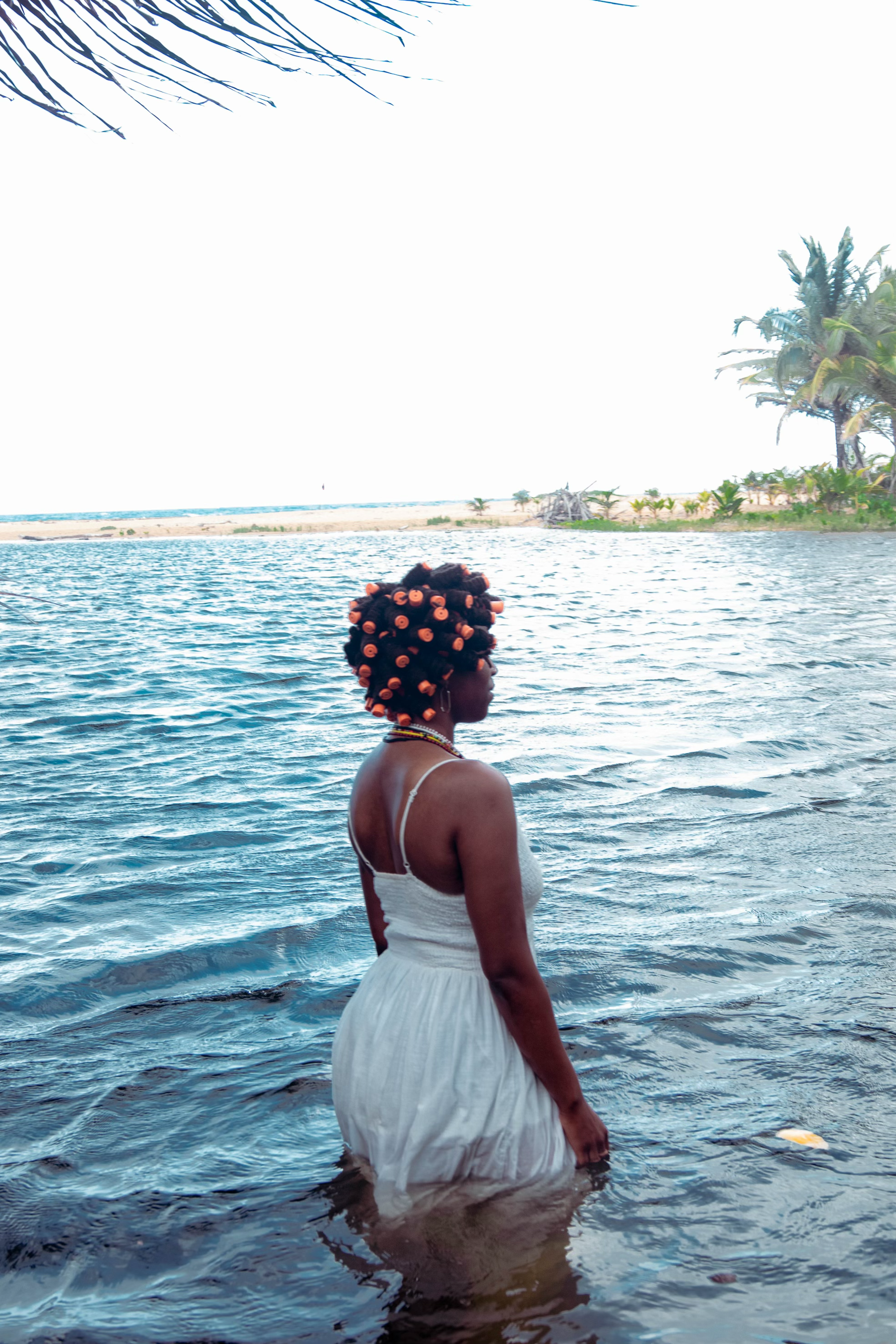 Déja Madkins in a white dress, standing in calm blue water beside a tropical shoreline.
